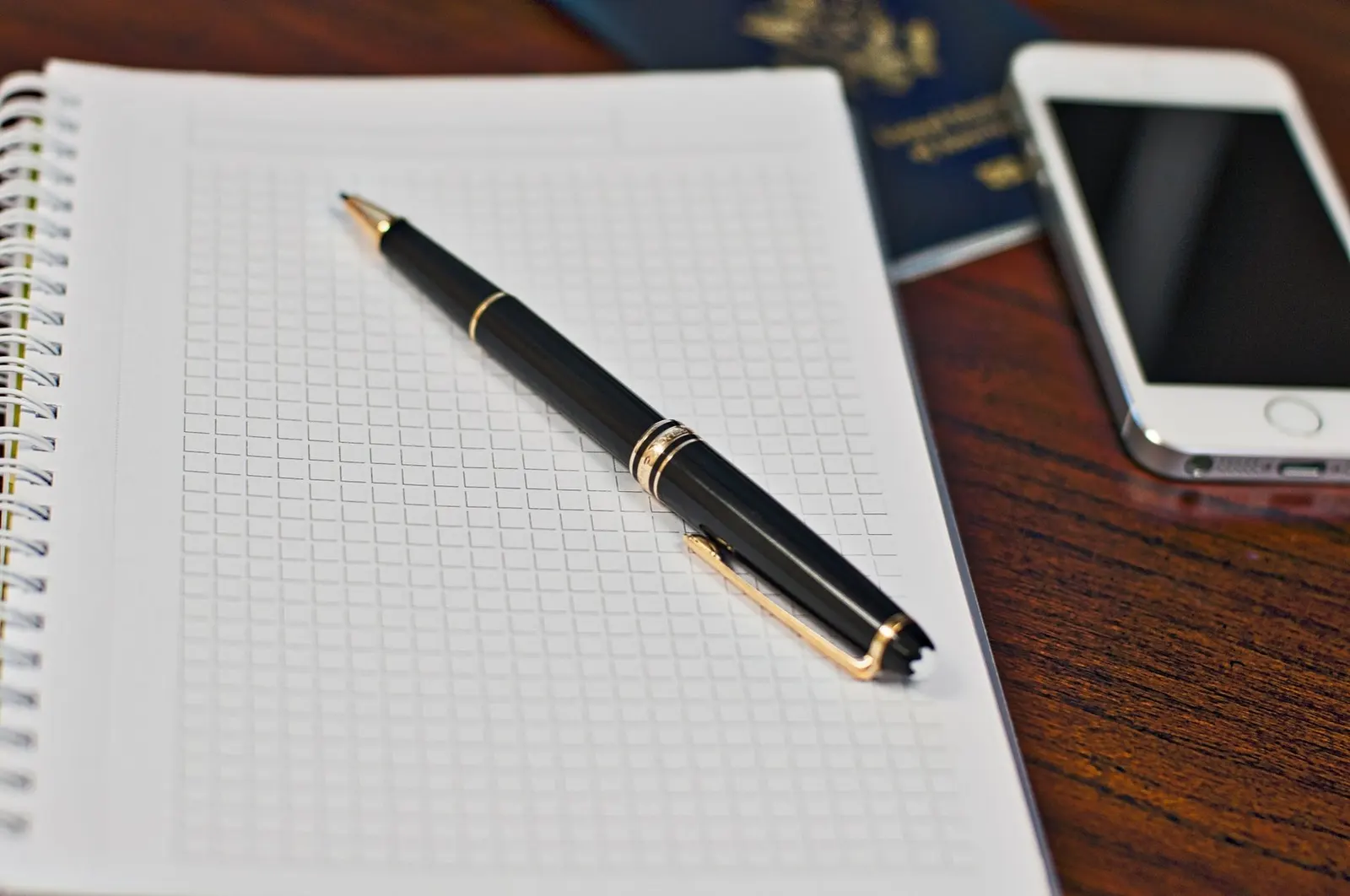 A closed passport and travel documents on a wooden desk beside a notebook and phone